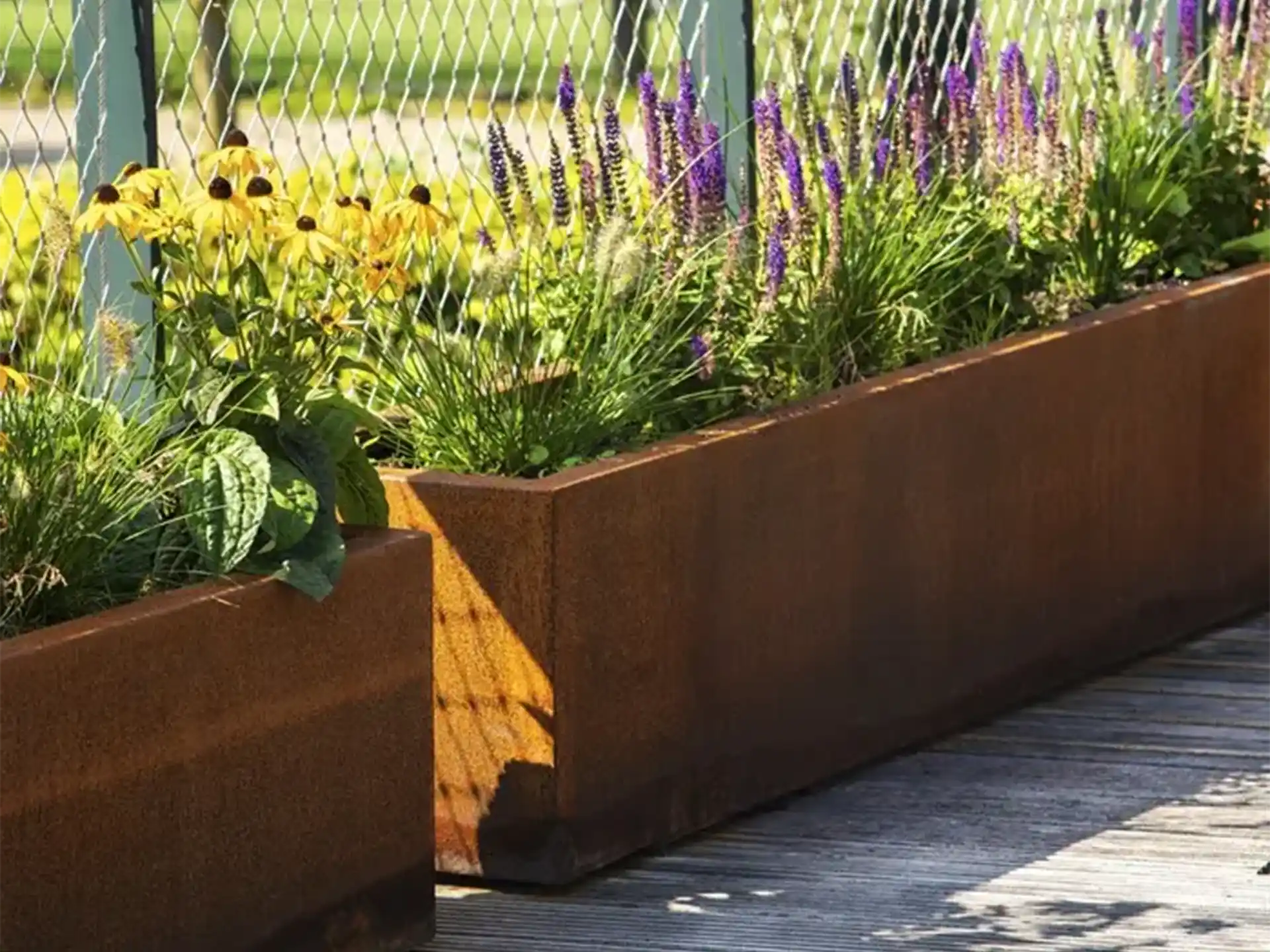 Grote rechthoekige Adezz Carrez Cortenstaal Plantenbak met wielen, beplant met bloemen en groen langs een moderne tuin.