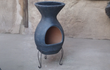 Sfeerfoto van de grijze Sol-y-Yo Barbecue terrashaard Acapulco (ø46 x 100 cm), in gebruik met brandend vuur en grillfunctie, sfeervol geplaatst in een tuinomgeving voor warmte en gezelligheid.