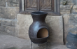 Sfeerfoto van de bruine Sol-y-Yo Chimenea Mexicaanse terracotta kachel Barbecue Jumbo (115 cm), brandend met open vuur en gebruikt als barbecue in een sfeervolle tuinomgeving.
