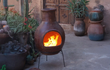 Sfeerfoto van de rode Sol-y-Yo Chimenea Mexicaanse terracotta kachel Jumbo (115 cm), brandend met open vuur in een sfeervolle tuinomgeving voor warmte en gezelligheid.