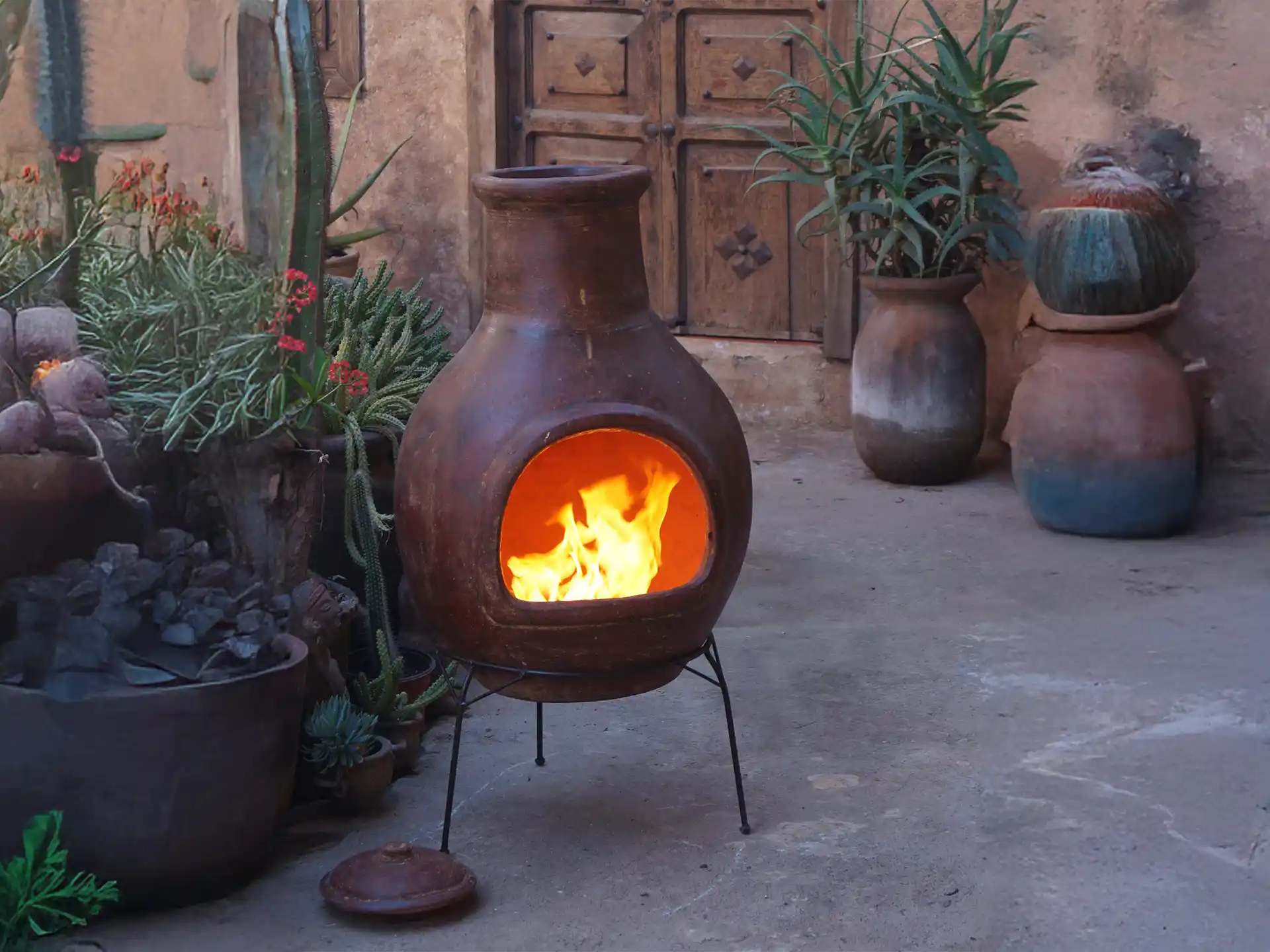 Sfeerfoto van de rode Sol-y-Yo Chimenea Mexicaanse terracotta kachel Jumbo (115 cm), brandend met open vuur in een sfeervolle tuinomgeving voor warmte en gezelligheid.
