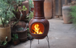 Sfeerfoto van de rode Sol-y-Yo Chimenea terracotta tuinhaard (105 cm) brandend in een tuin, gebruikt als buitenhaard voor warmte en gezelligheid.