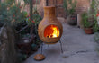 Sfeerfoto van de gele Sol-y-Yo Mexicaanse terracotta kachel Jumbo (115 cm), brandend met open vuur in een sfeervolle tuinomgeving voor warmte en gezelligheid.