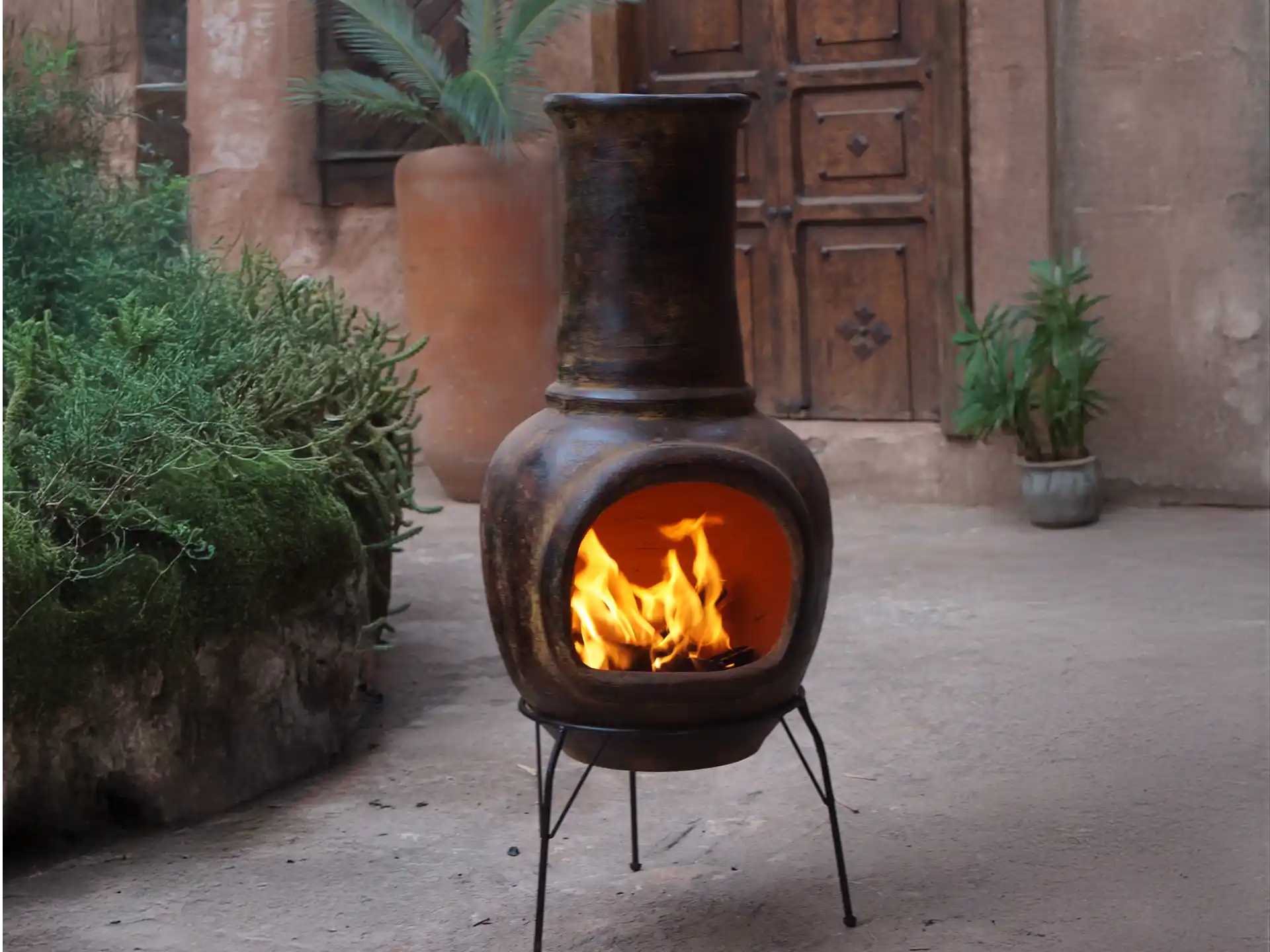 Sfeerfoto van de grijze Sol-y-Yo Mexicaanse terrashaard (105 cm) met voet en deksel, brandend in een tuinomgeving voor warmte en gezelligheid.