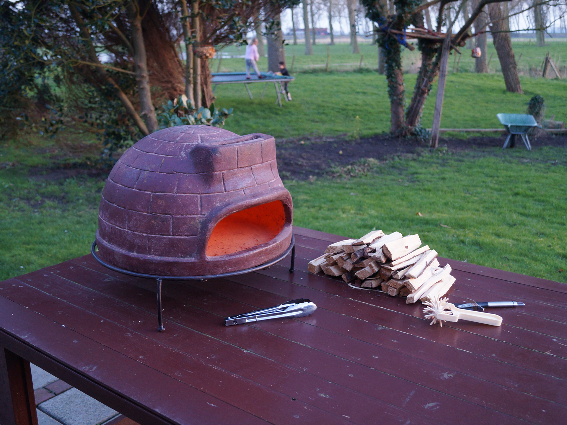 Sol-y-Yo Venetiaanse houtgestookte pizza oven 52 cm, buiten geplaatst op houten tafel, klaar voor gebruik met houtblokken ernaast.