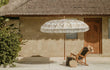 Crème Bali parasol met goudkleurige beschildering en franjes, geplaatst bij een rotan loungeset in een zonnige tuin.