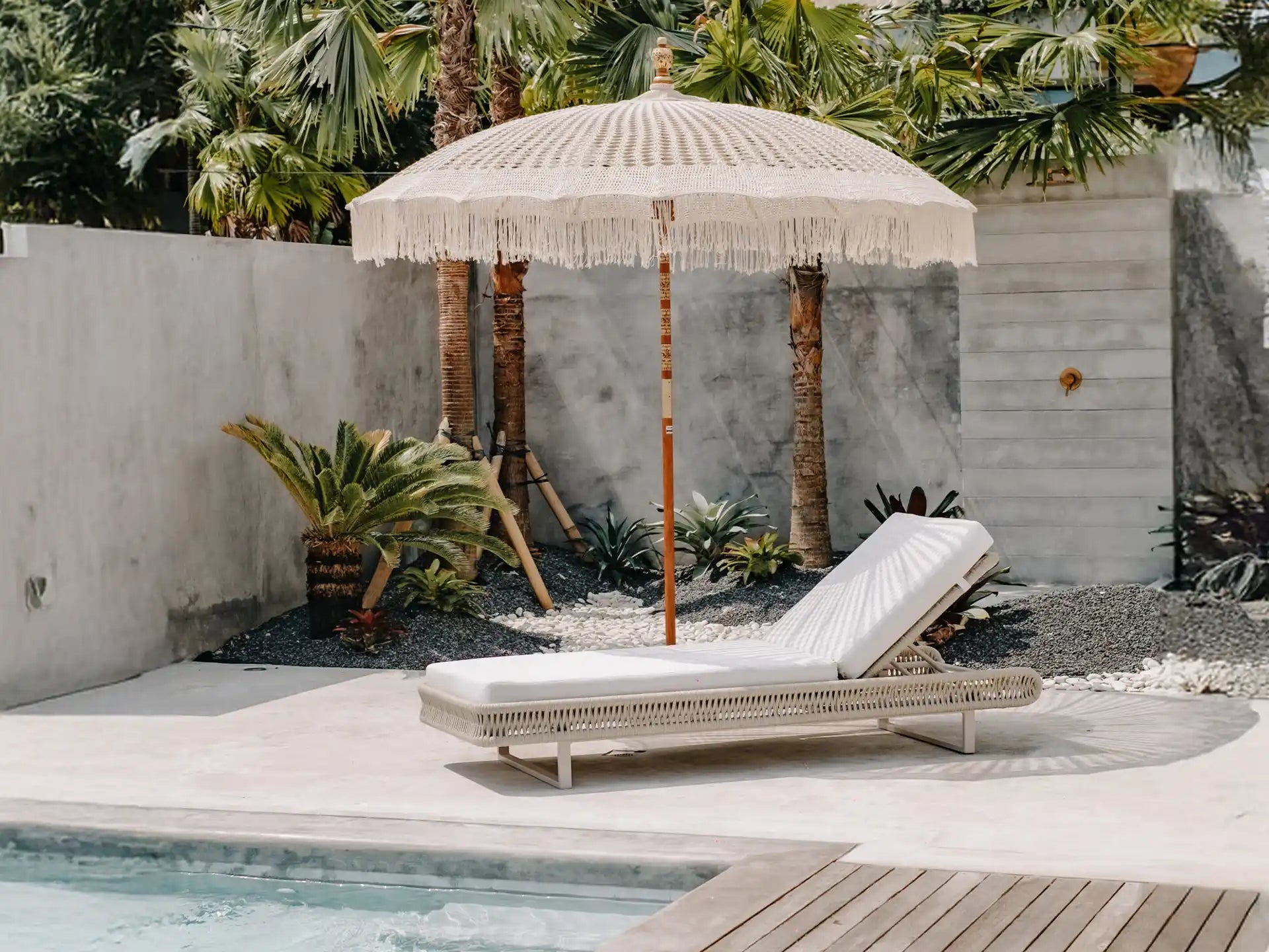 Stijlvolle crème Bali parasol met macramé details en franjes, geplaatst naast een modern zwembad met tropische planten en loungebed, perfect voor een bohemian tuinsetting.