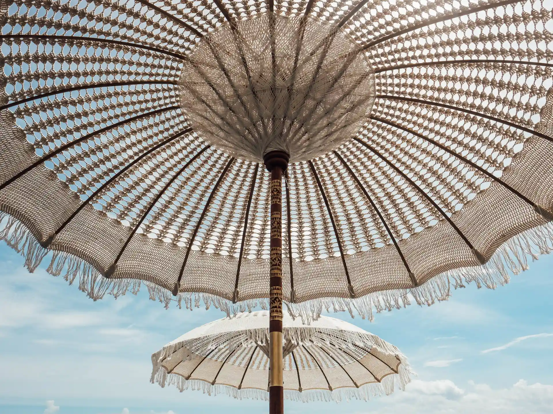 Detailopname van een crème Bali parasol met macramé franjes, gezien van onderaf tegen een heldere blauwe lucht, sfeervol bohemian design voor tuin of terras.