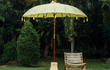 Gele Todo Bien Bali parasol in een tropische tuinsetting naast een houten stoel, ideaal voor zomerse loungesfeer.