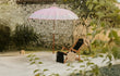 Bali parasol van Todo Bien in roze met zilveren rand, opgesteld naast een rotan stoel bij een zwembad in een tropische tuin.