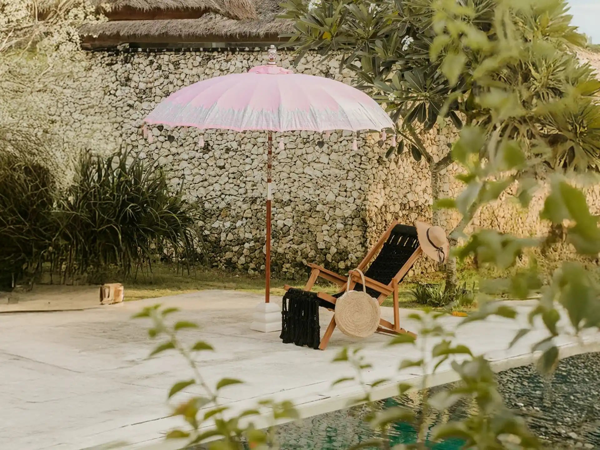 Bali parasol van Todo Bien in roze met zilveren rand, opgesteld naast een rotan stoel bij een zwembad in een tropische tuin.