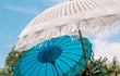 Detail van een handgemaakte Bali parasol in zeeblauw met witte macramé franjes en houten stok, gefotografeerd tegen een heldere blauwe lucht.