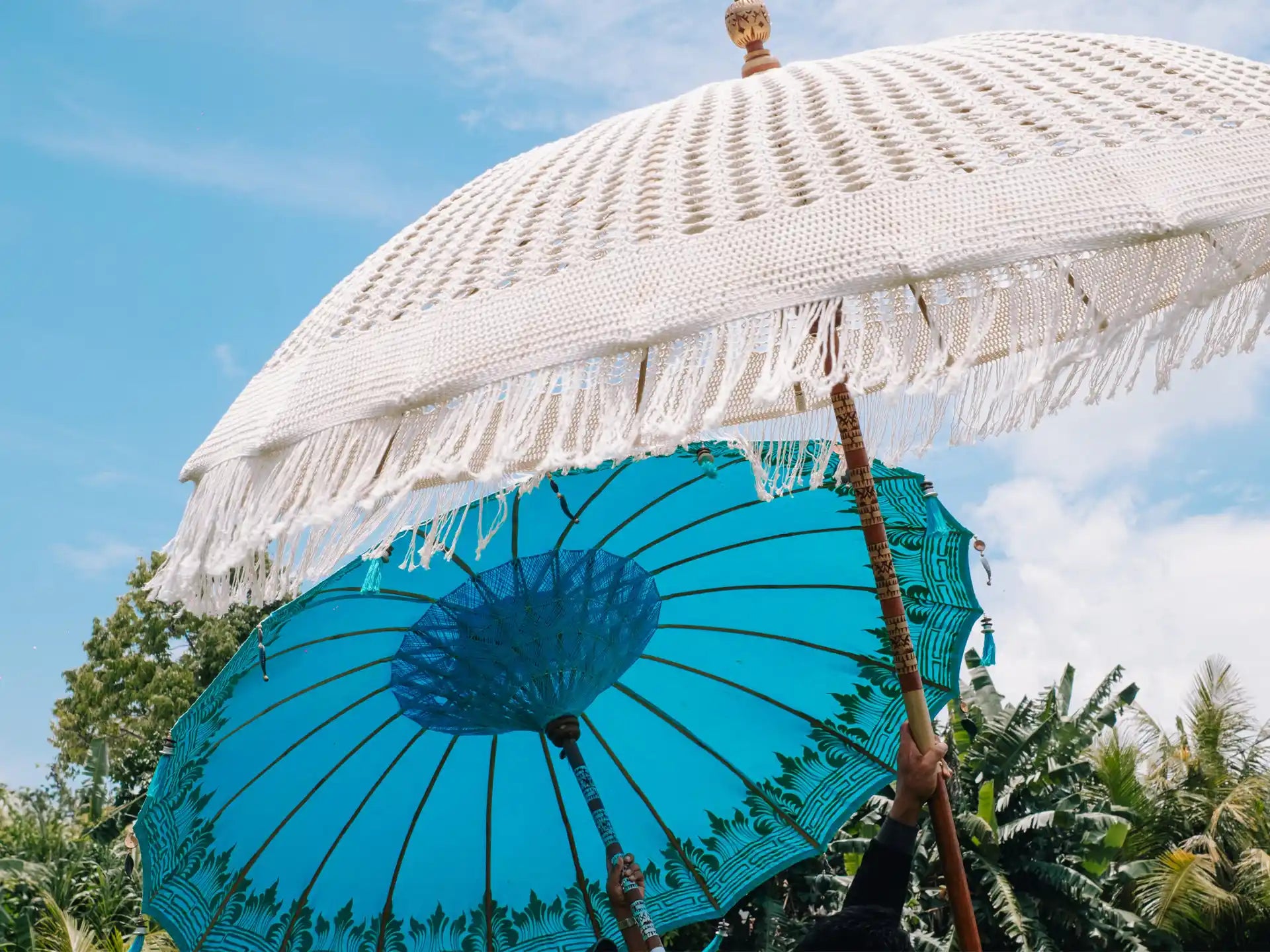 Detail van een handgemaakte Bali parasol in zeeblauw met witte macramé franjes en houten stok, gefotografeerd tegen een heldere blauwe lucht.