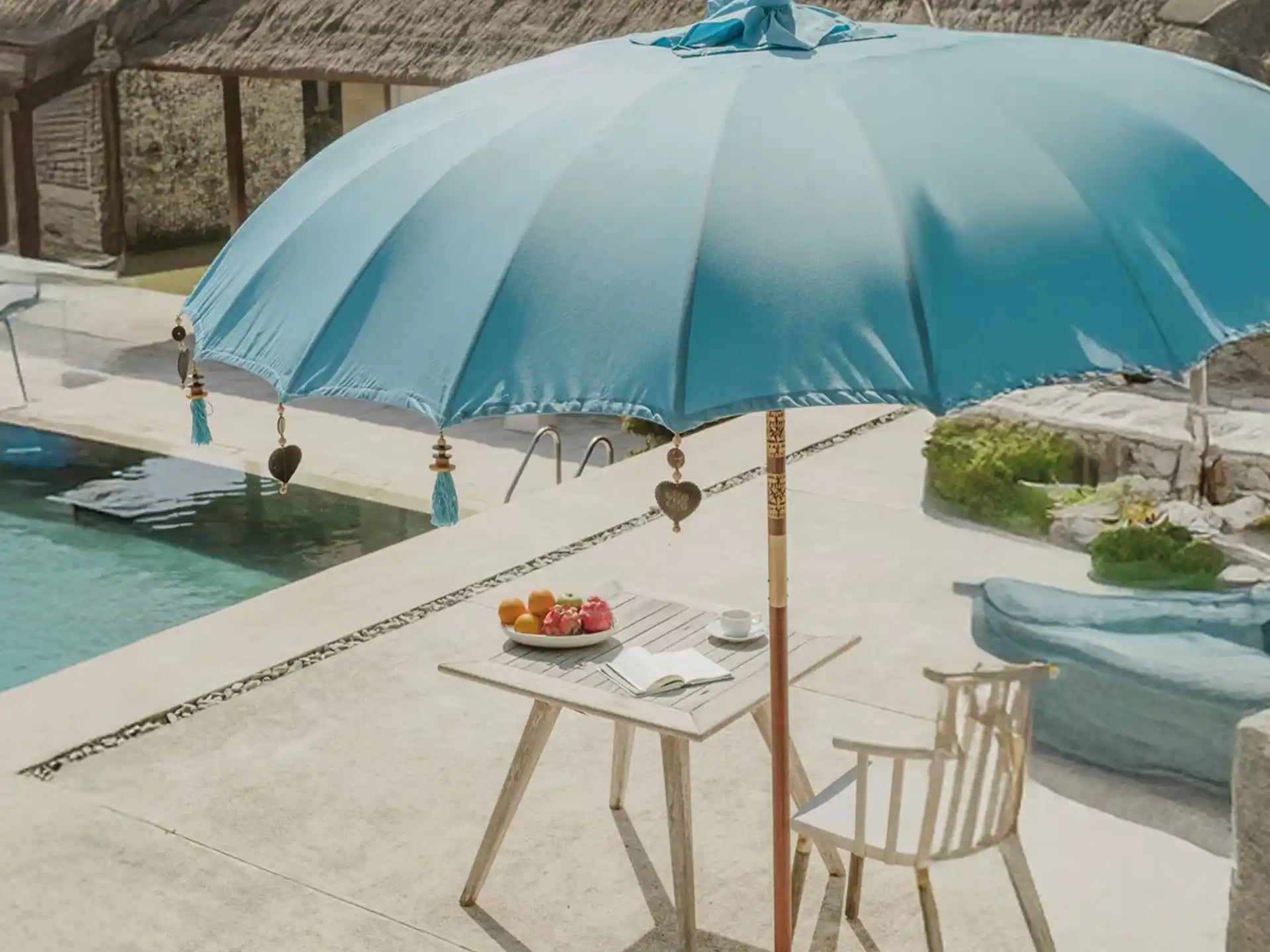 Sfeerfoto van de Todo Bien Bali parasol zeeblauw bij een zwembad, met houten tafel, fruit en stoel voor een zomerse luxe uitstraling.