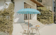 Sfeervolle tuinopstelling met de Todo Bien Bali parasol in zeeblauw, geplaatst naast een wit huis en houten tafel, ideaal voor schaduw en luxe uitstraling in de tuin.