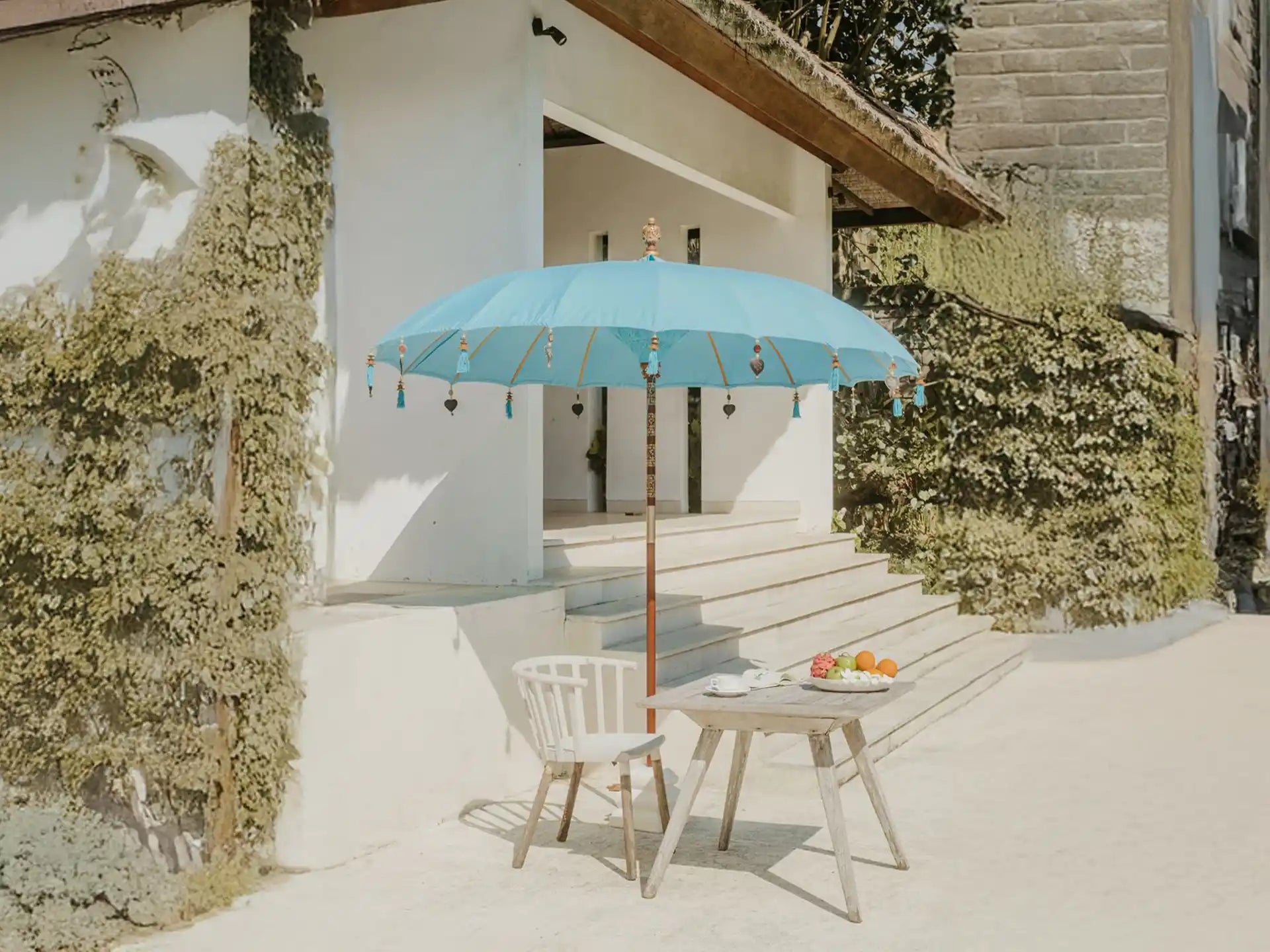Sfeervolle tuinopstelling met de Todo Bien Bali parasol in zeeblauw, geplaatst naast een wit huis en houten tafel, ideaal voor schaduw en luxe uitstraling in de tuin.