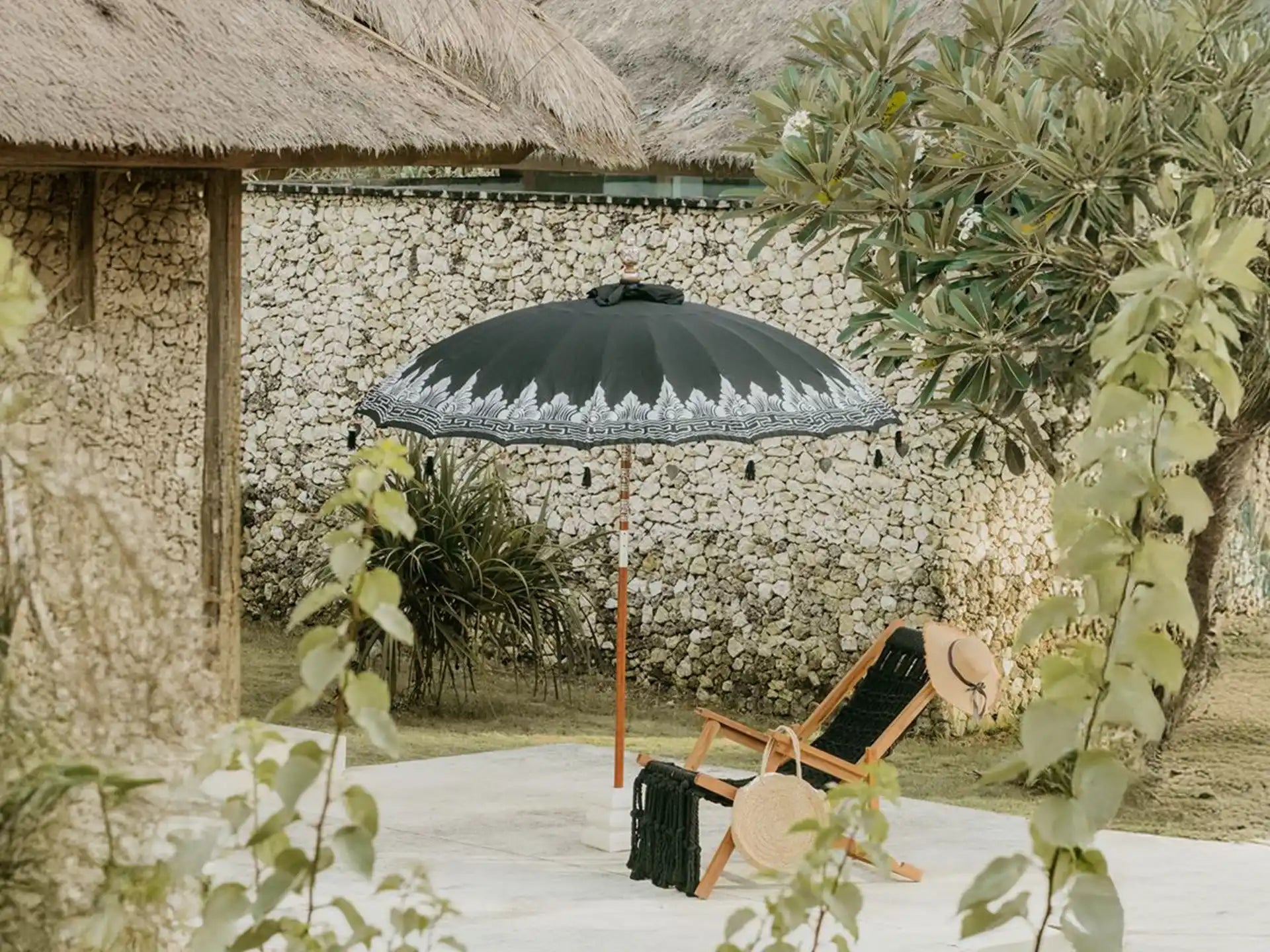 Sfeervolle opstelling van de zwarte Bali parasol met zilveren beschildering bij een ligstoel naast een stenen muur.