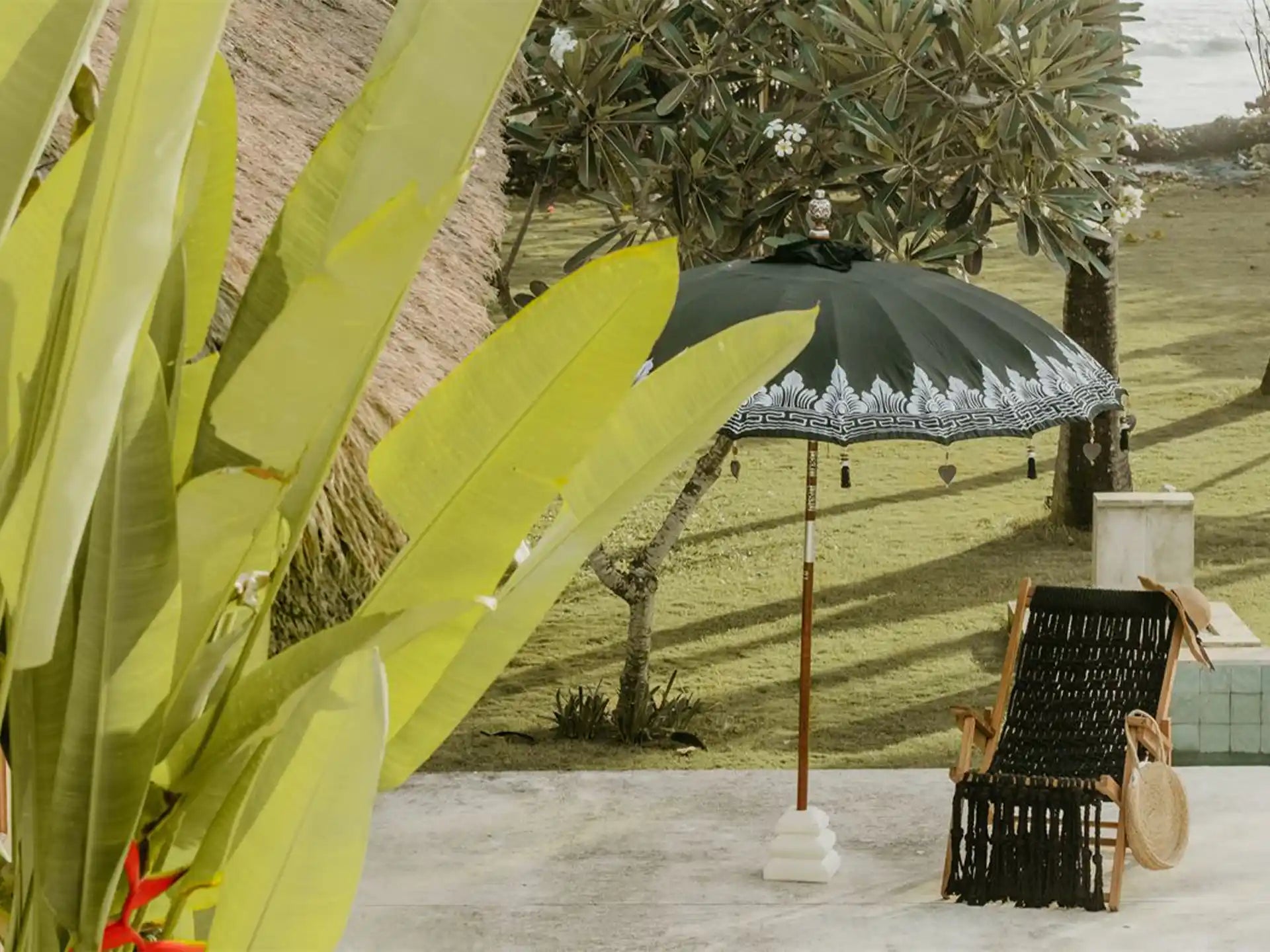 Zwarte Bali parasol met zilveren beschildering geplaatst in een tropische tuin naast een ligstoel en planten.